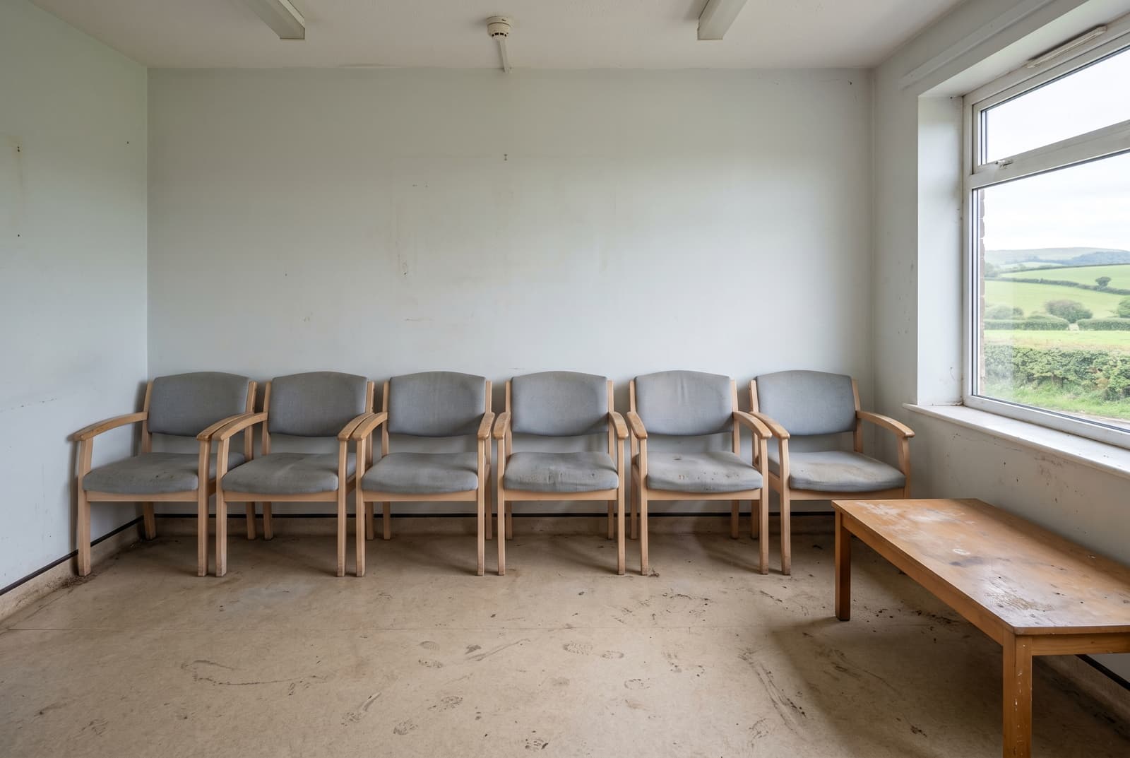 Devon GP surgery waiting room before cleaning, scuffed hazy vinyl floor, dusty chairs and marked low table